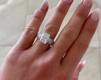 Può includere: Un anello di diamanti con una pietra centrale taglio rettangolare e una fascia pavé. L'anello presenta un grande diamante rettangolare, circondato da diamanti più piccoli. La fascia è anch'essa incastonata con piccoli diamanti, creando un effetto scintillante.