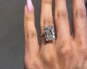 Può includere: Un anello di fidanzamento in oro con un grande diamante rettangolare trasparente. L'anello è su un dito.