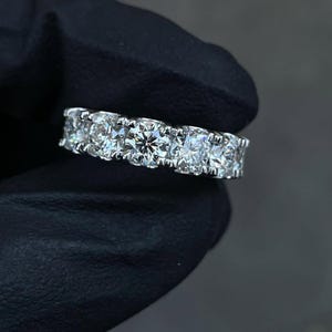 May include: A close-up of a silver ring with multiple round-cut diamonds. The diamonds are set closely together, creating a continuous band of sparkle. The ring is held against a dark background.