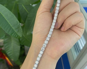 May include: A tennis bracelet featuring a line of round, brilliant-cut diamonds. The bracelet is set in a silver or white gold metal and is held against a background of green foliage.