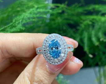 May include: A silver ring with a large, oval-shaped, light blue gemstone, encircled by two halos of smaller, clear gemstones. The ring's band is also set with small, clear gemstones. The ring is displayed against a blurred green backdrop.