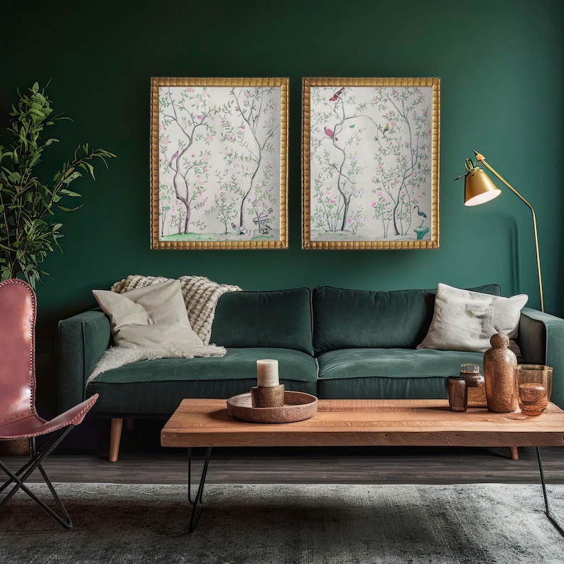 The listing’s pair of antique chinoiserie prints of wispy trees with green leaves and pastel-colored blossoms hosting various colorful birds and a butterfly against an off-white background hangs in gold frames on a deep green wall above a green velvet sofa with pillows, a wooden coffee table with a tray and a candle, a gold floor lamp and a potted plant.
