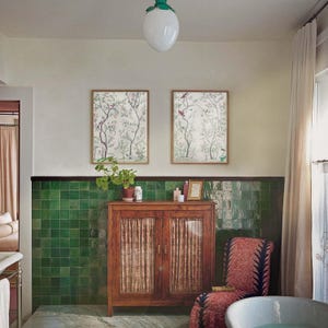The listing’s pair of antique chinoiserie prints of wispy trees with green leaves and pastel-colored blossoms hosting various colorful birds and a butterfly against an off-white background hangs on the wall of a bohemian bathroom with green tile wainscoting, a white clawfoot bathtub, a cozy world-market chair and a vintage wood cabinet.