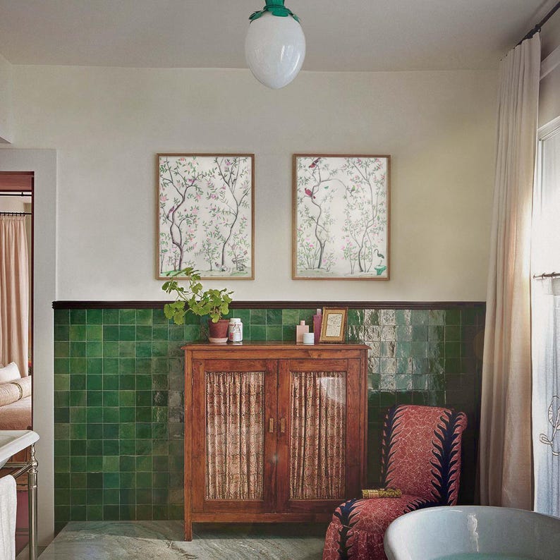 The listing’s pair of antique chinoiserie prints of wispy trees with green leaves and pastel-colored blossoms hosting various colorful birds and a butterfly against an off-white background hangs on the wall of a bohemian bathroom with green tile wainscoting, a white clawfoot bathtub, a cozy world-market chair and a vintage wood cabinet.