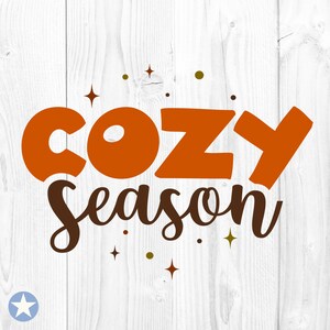 Może przedstawiać: Pomarańczowy i brązowy tekst na białym drewnianym tle, który brzmi "Cozy Season". Tekst jest otoczony małymi złotymi i białymi gwiazdkami.