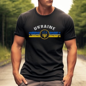 May include: A black t-shirt with a distressed yellow and blue stripe design and the word "UKRAINE" in white lettering. A yellow circle with a blue trident symbol is centered on the stripe design.