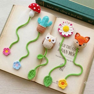 May include: Five handmade crochet bookmarks featuring a red mushroom, a green cactus in a brown pot, a white owl, a white daisy with a yellow center, and an orange fox. Each bookmark has a long green stem ending in a small crocheted flower or leaf.