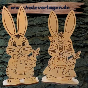 Op de afbeelding: Twee houten paashaasfiguren, een mannetje en een vrouwtje, die elk een wortel vasthouden. Het mannetje draagt een vlinderdas en het vrouwtje een strik op haar hoofd. De figuren zijn lichtbruin met geëtste details. De achtergrond is een getextureerd, donkergroen oppervlak.