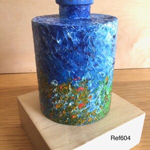 May include: A blue and green hand-painted ceramic vase with a floral design. The vase has a textured surface and a round shape. The vase is sitting on a wooden base with the text "Ref604" printed on it.