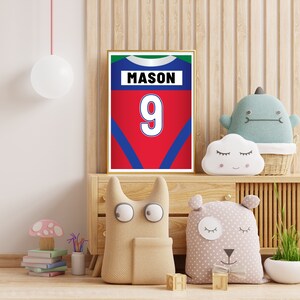 Puede incluir: Una impresi&oacute;n enmarcada de una camiseta roja, azul y verde con el nombre "MASON" y el n&uacute;mero "9" en ella. La impresi&oacute;n est&aacute; colgada en una pared en una habitaci&oacute;n con una c&oacute;moda de madera y juguetes de peluche.