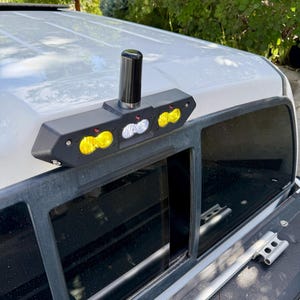 May include: A black, angular, third brake light housing mounted on the roof of a white vehicle, featuring two yellow LED light pods on each side and a central white LED light pod, topped with a vertical, cylindrical black antenna.
