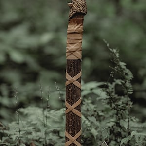 May include: A hand-crafted axe with a wooden handle wrapped in brown leather. The handle is carved with a wolf's head at the top. The axe head is black and has a serrated edge.