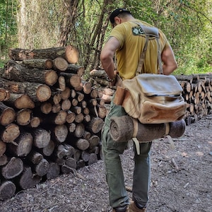 May include: A person wearing a yellow t-shirt with a patch on the back, green cargo pants, and a brown leather backpack with a rolled-up blanket strapped to the side. The person is standing in a forest setting with a pile of logs in the background.