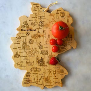 Könnte beinhalten: Ein Holzbrett in Form Deutschlands mit einer eingravierten Karte des Landes und wichtigen Städten. Das Brett ist mit einer roten Tomate, drei kleinen roten Tomaten und einer Erdbeere verziert.
