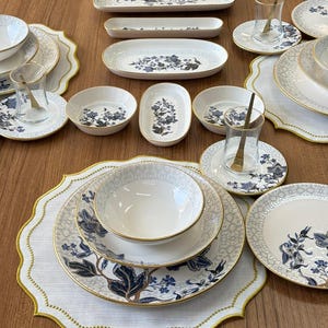 Op de afbeelding: Een uitgebreide serviesset bestaande uit witte keramische borden, kommen en serveerschalen versierd met blauwe bloemmotieven en goudkleurige randen. De collectie bevat bijpassende glazen theekopjes op schoteltjes en geschulpte placemats.