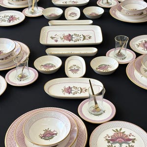 Op de afbeelding: Een servies bestaande uit witte keramische borden, kommen en schalen met een roze en groen bloemmotief en een gouden rand. De set bevat kleine theeglazen met gouden lepels, gerangschikt op een zwarte ondergrond voor een complete tafelsetting.