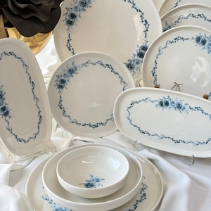 May include: A collection of white ceramic dinnerware featuring a delicate blue floral pattern. The set includes round plates, oval platters, and bowls, each decorated with a wreath-like arrangement of blue roses and small leaves along the edges.