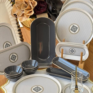 May include: A collection of ceramic dinnerware featuring white plates with black lattice patterns and central floral medallions, dark grey rectangular serving platters, small grey bowls, and a clear glass tea cup with a gold spoon on a matching saucer.