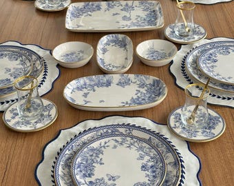 May include: A full dinner service with white and blue floral designs. The set includes plates, bowls, serving dishes, and teacups with gold rims. White placemats with a blue scalloped edge are also included. The table setting is arranged on a wooden surface.