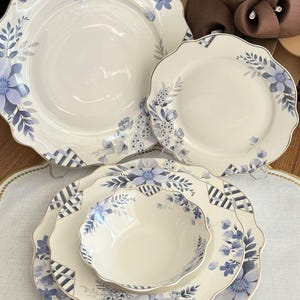 Puede incluir: Un juego de vajilla de porcelana color crema con un diseño floral azul. El juego incluye platos y un cuenco con bordes festoneados y un patrón geométrico. El patrón floral presenta flores y hojas azules.