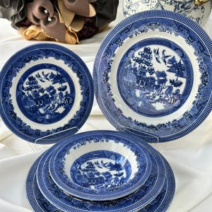 Puede incluir: Un conjunto de vajilla de cerámica azul y blanca con el clásico patrón Willow. El juego incluye platos y cuencos de varios tamaños, con un diseño detallado de paisaje. La vajilla está dispuesta sobre una superficie blanca, mostrando la vajilla tradicional.