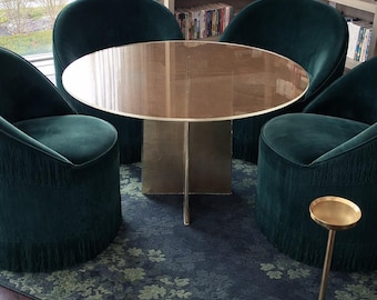 Puede incluir: Una mesa de comedor redonda con tablero de cristal reflectante y base metálica, rodeada de cuatro sillas de terciopelo verde oscuro con flecos. Una pequeña mesa auxiliar dorada se encuentra cerca sobre una alfombra con estampado floral.