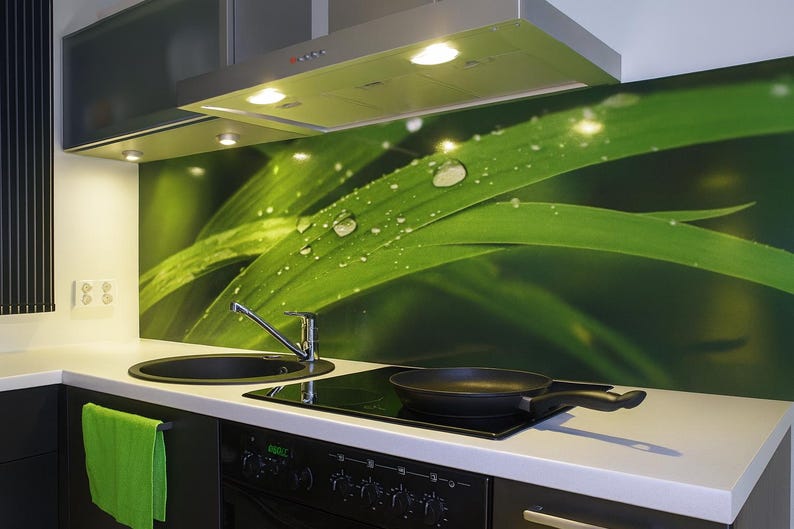 Puede incluir: Una cocina moderna con vitrocer&aacute;mica, fregadero y campana extractora negros. Un protector contra salpicaduras verde presenta un primer plano de hojas verdes con gotas de agua. Una sart&eacute;n negra descansa sobre la vitrocer&aacute;mica y una toalla verde cuelga.