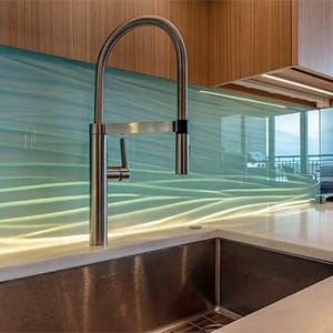 Puede incluir: Una cocina moderna con un grifo y un fregadero de acero inoxidable. El protector contra salpicaduras tiene un dise&ntilde;o ondulado en azul claro y verde. La encimera es blanca y los armarios son de madera.