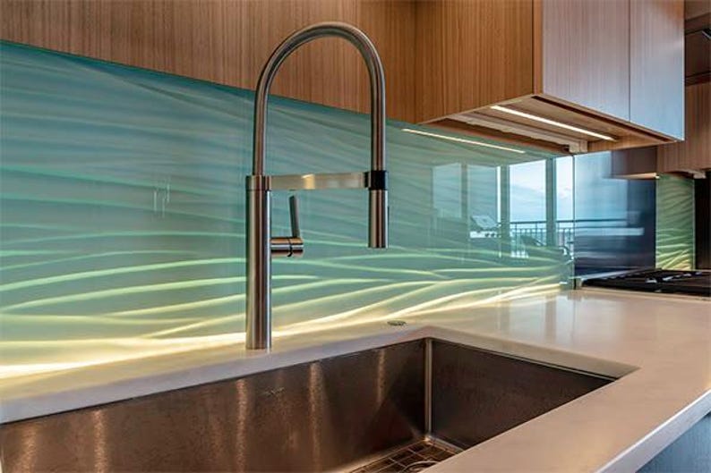 Puede incluir: Una cocina moderna con un grifo y un fregadero de acero inoxidable. El protector contra salpicaduras tiene un dise&ntilde;o ondulado en azul claro y verde. La encimera es blanca y los armarios son de madera.