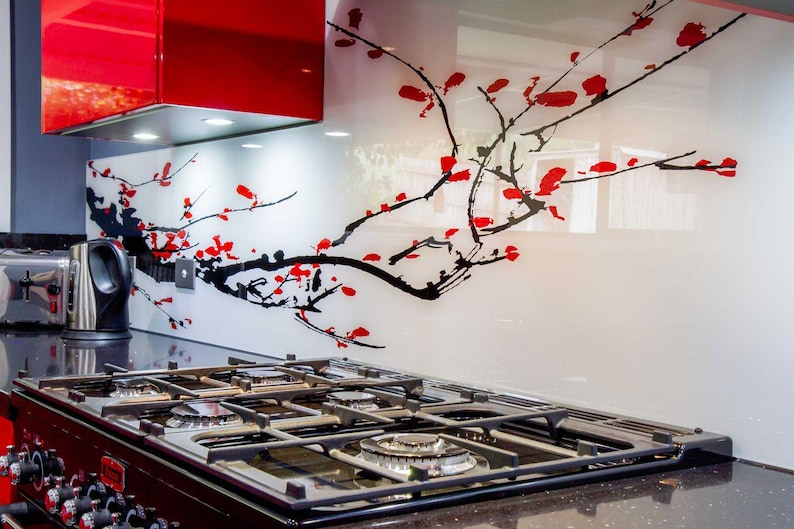 Puede incluir: Una cocina moderna con una estufa de gas negra, una campana extractora roja y un protector contra salpicaduras blanco con un dise&ntilde;o floral negro y rojo. Una tostadora y una tetera de acero inoxidable est&aacute;n en la encimera.