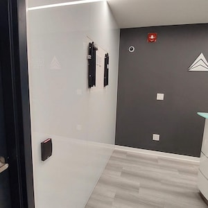 Puede incluir: Vista interior de una oficina moderna con paredes blancas y grises, y suelo gris claro con aspecto de madera. Un marco de puerta negro es visible a la izquierda. Las paredes tienen un dise&ntilde;o minimalista con un panel de acceso de seguridad y un soporte de montaje.