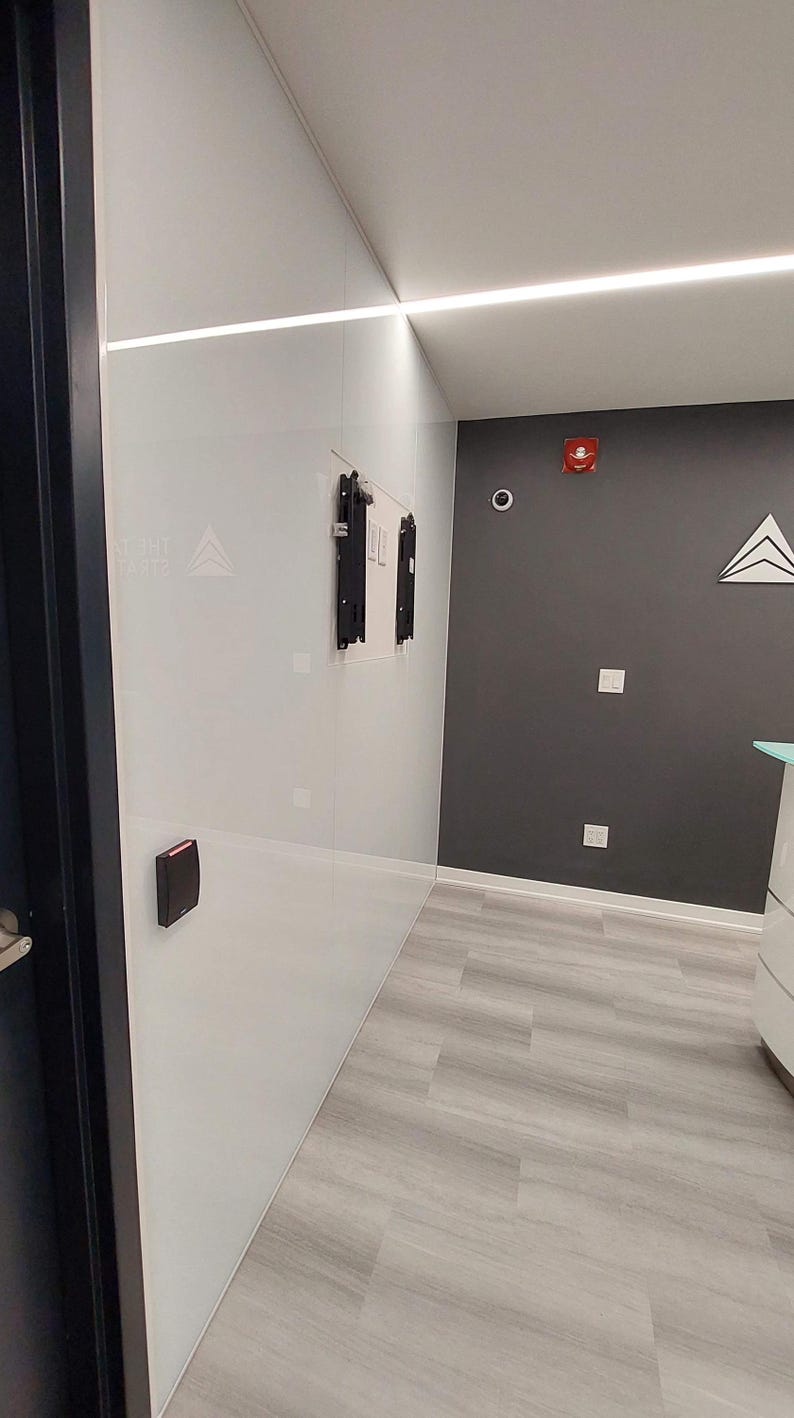 Puede incluir: Vista interior de una oficina moderna con paredes blancas y grises, y suelo gris claro con aspecto de madera. Un marco de puerta negro es visible a la izquierda. Las paredes tienen un dise&ntilde;o minimalista con un panel de acceso de seguridad y un soporte de montaje.