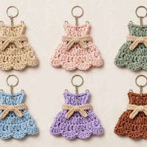 May include: A set of six crochet dress keychains in beige, light pink, sage green, light blue, lavender, and brown. Each dress features a scalloped hem, a fitted bodice, and a small beige crocheted bow at the waist, attached to a silver metal ring.