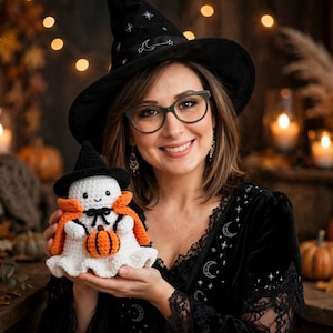 Könnte beinhalten: Eine weiß gehäkelte Geisterfigur mit einem schwarzen spitzen Hut und einem orangefarbenen Umhang, die einen kleinen orange-schwarzen gehäkelten Kürbis hält. Der Geist hat schwarze gestickte Augen und ein kleines Lächeln aus weichem Garn.