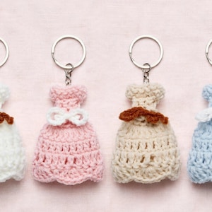 Peut inclure: Quatre porte-clés en forme de robe au crochet alignés. Les robes sont blanches, rose clair, beiges et bleu clair. Chaque robe présente un petit nœud crocheté à la taille, soit marron soit blanc, et est fixée à un anneau porte-clés en métal argenté.