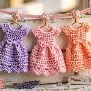 May include: Three miniature crochet dresses in lavender, light pink, and peach, each hanging on a gold wire hanger with a small flower and leaf accent. The dresses feature a short-sleeve bodice, a tied waist bow, and a scalloped hemline.