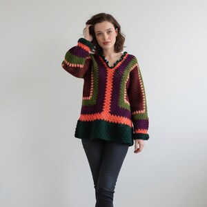 Peut inclure: Un pull en crochet à manches longues doté d'une encolure en V avec un bord festonné. Le vêtement est composé de blocs de laine multicolores dans des tons vert foncé, violet profond et orange vif, disposés en un motif géométrique.
