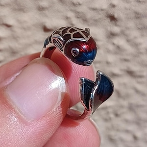 Könnte beinhalten: Ein silberner Ring mit Fischdesign. Der Fisch ist mit roter, blauer und brauner Emaille verziert. Der Schwanz des Rings umschließt den Finger und schafft ein einzigartiges Accessoire. Der Ring ist ein Schmuckstück.