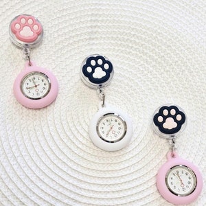 May include: Three pink and white fob watches with paw print charms. The watches have black hands and black numbers. The charms are attached to a retractable clip.