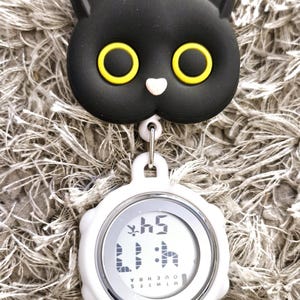 Peut inclure: Un clip en forme de chat noir avec des yeux jaunes attach&eacute; &agrave; un minuteur num&eacute;rique blanc avec un bord argent&eacute;. Le minuteur affiche "1:15" et les mots "MINUTES" et "HEURES" sont visibles.