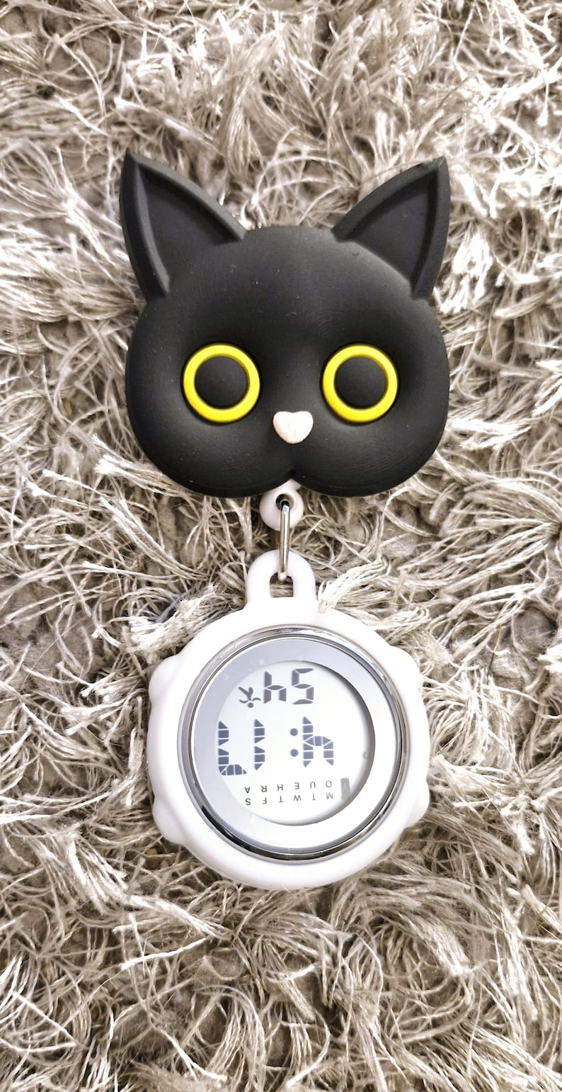 Peut inclure: Un clip en forme de chat noir avec des yeux jaunes attach&eacute; &agrave; un minuteur num&eacute;rique blanc avec un bord argent&eacute;. Le minuteur affiche "1:15" et les mots "MINUTES" et "HEURES" sont visibles.