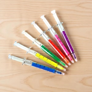 Op de afbeelding: Zes kleurrijke pennen in de vorm van een spuit, diagonaal gerangschikt op een licht houten oppervlak. De pennen hebben doorzichtige cilinders gevuld met blauwe, gele, groene, rode, roze en paarse inkt. Elke pen heeft een witte zuiger en een transparante punt.