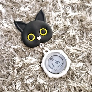 Peut inclure: Un porte-badge r&eacute;tractable en forme de chat noir avec des yeux jaunes et une montre num&eacute;rique blanche attach&eacute;e.