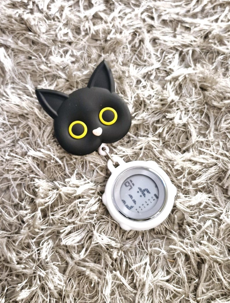 Peut inclure: Un porte-badge r&eacute;tractable en forme de chat noir avec des yeux jaunes et une montre num&eacute;rique blanche attach&eacute;e.