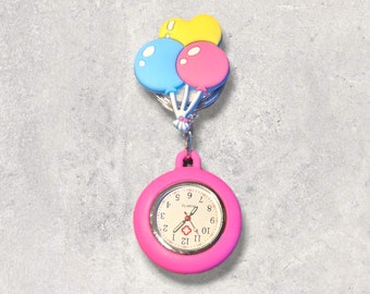Puede incluir: Un reloj de bolsillo rosa con una esfera blanca y agujas negras. El reloj está unido a un charm con tres globos de colores: amarillo, azul y rosa.