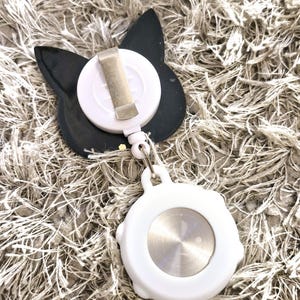 Peut inclure: Un porte-badge r&eacute;tractable en forme de chat noir et blanc avec un clip argent&eacute;. Le porte-badge est attach&eacute; &agrave; une bobine r&eacute;tractable blanche avec un bouton argent&eacute;.