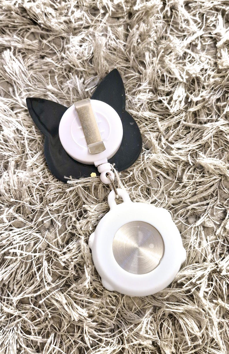 Peut inclure: Un porte-badge r&eacute;tractable en forme de chat noir et blanc avec un clip argent&eacute;. Le porte-badge est attach&eacute; &agrave; une bobine r&eacute;tractable blanche avec un bouton argent&eacute;.