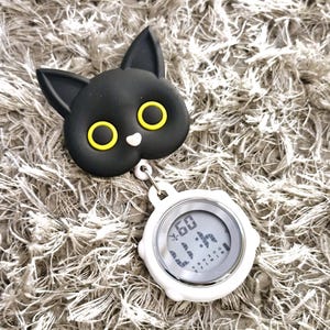 Peut inclure: Un clip en forme de chat noir avec des yeux jaunes attach&eacute; &agrave; un chronom&egrave;tre num&eacute;rique blanc avec un cadran noir. Le chronom&egrave;tre affiche "60" et "1:14" en police num&eacute;rique.