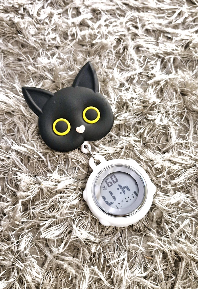 Peut inclure: Un clip en forme de chat noir avec des yeux jaunes attach&eacute; &agrave; un chronom&egrave;tre num&eacute;rique blanc avec un cadran noir. Le chronom&egrave;tre affiche "60" et "1:14" en police num&eacute;rique.