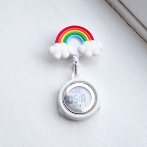 Op de afbeelding: Een wit digitaal zusterhorloge met een kleurrijke regenboog en wolken als opzetstuk. De ronde wijzerplaat heeft een zilverkleurige rand en een digitaal display dat de tijd weergeeft. Het horloge is voorzien van een clip voor aan de kleding.